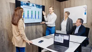 A person presents a bar chart on a screen to three colleagues in a meeting room with laptops and a whiteboard.
