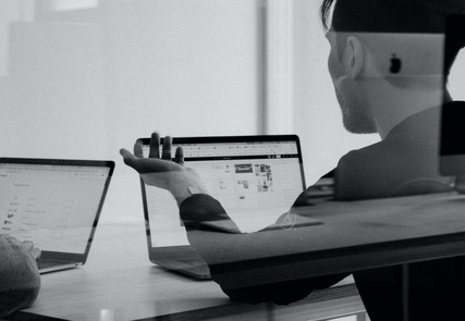 Person gesturing while discussing something on a laptop screen with another individual, in a modern office setting. Black and white image.