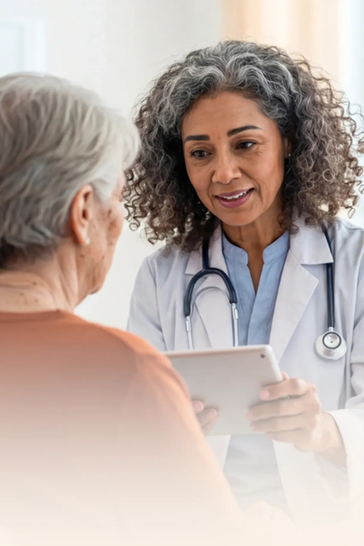 Doctor with curly hair and stethoscope holds a tablet, attentively conversing with an elderly woman in a brown shirt.