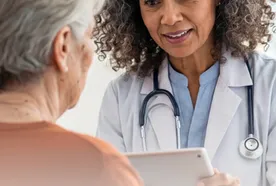 Doctor with curly hair and stethoscope holds a tablet, attentively conversing with an elderly woman in a brown shirt.
