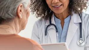 Doctor with curly hair and stethoscope holds a tablet, attentively conversing with an elderly woman in a brown shirt.