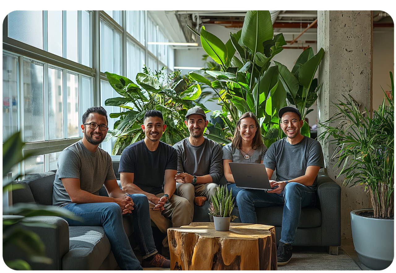 Five people sitting on sofas in a modern office with large windows and lush green plants, smiling and holding a laptop.