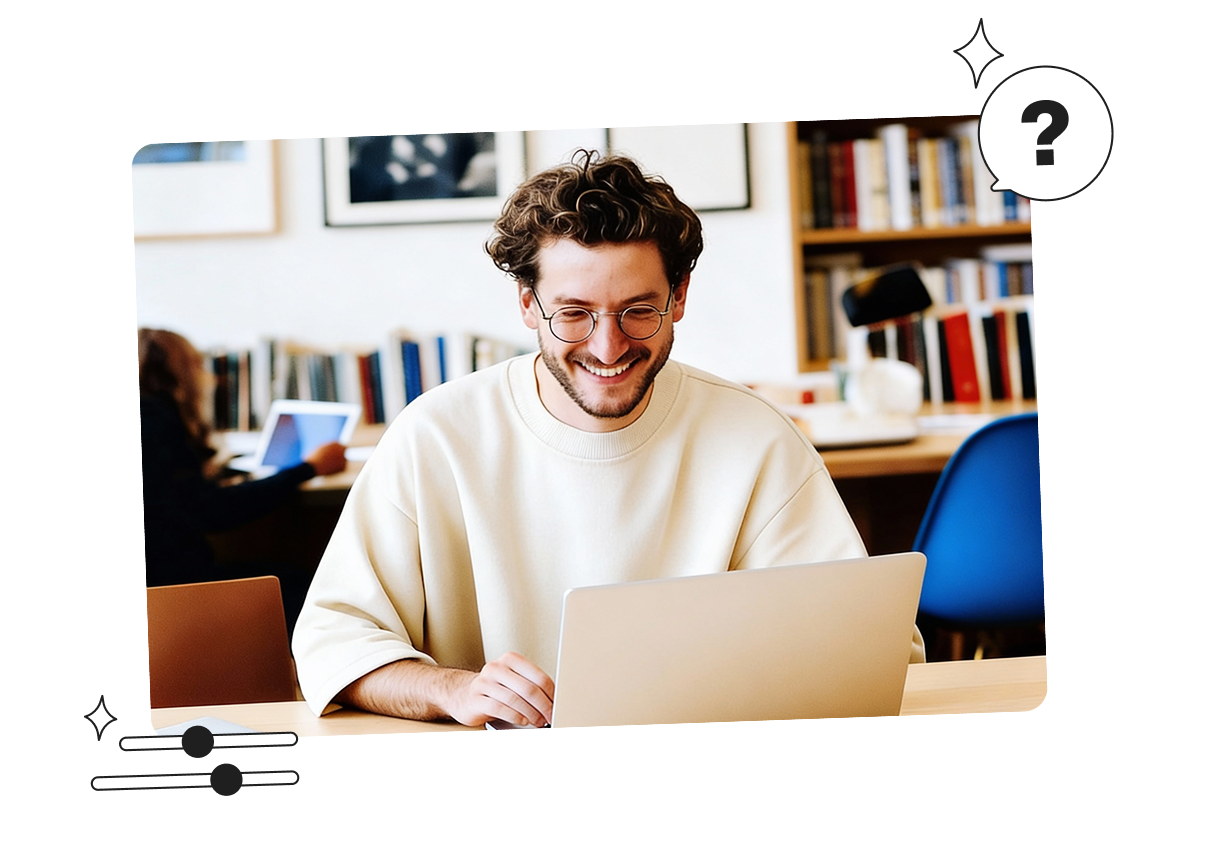A person with curly hair and glasses is smiling while using a laptop in a library setting. Another person is in the background with bookshelves.