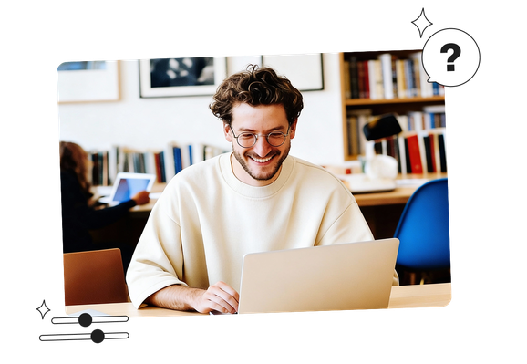 A person with curly hair and glasses is smiling while using a laptop in a library setting. Another person is in the background with bookshelves.