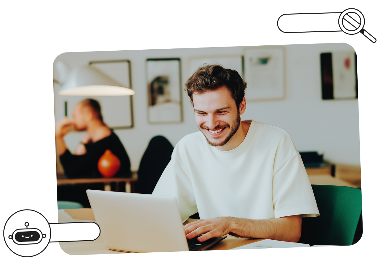Smiling man in a white shirt using a laptop at a desk in a modern office, with blurred coworker in the background.