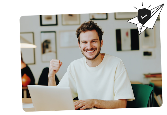 Smiling man in a white shirt sitting at a laptop, raising a fist in success. Background features a gallery wall and a paper plane graphic.
