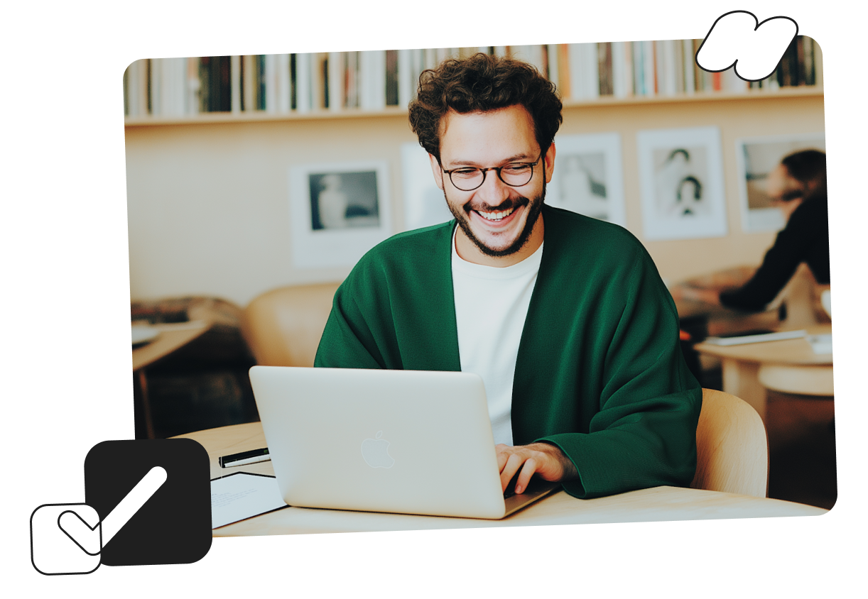 Smiling man in a green sweater using a laptop in a cozy library with bookshelves in the background.