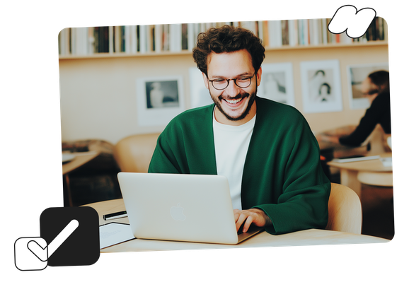 Smiling man in a green sweater using a laptop in a cozy library with bookshelves in the background.