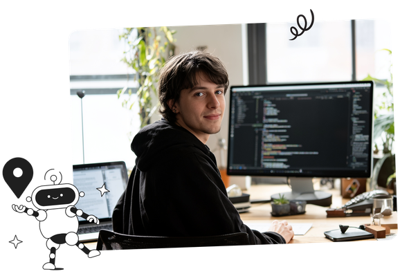 A person in a black hoodie sits at a desk with dual monitors displaying code, looking over their shoulder. Office plants and a window are in the background.