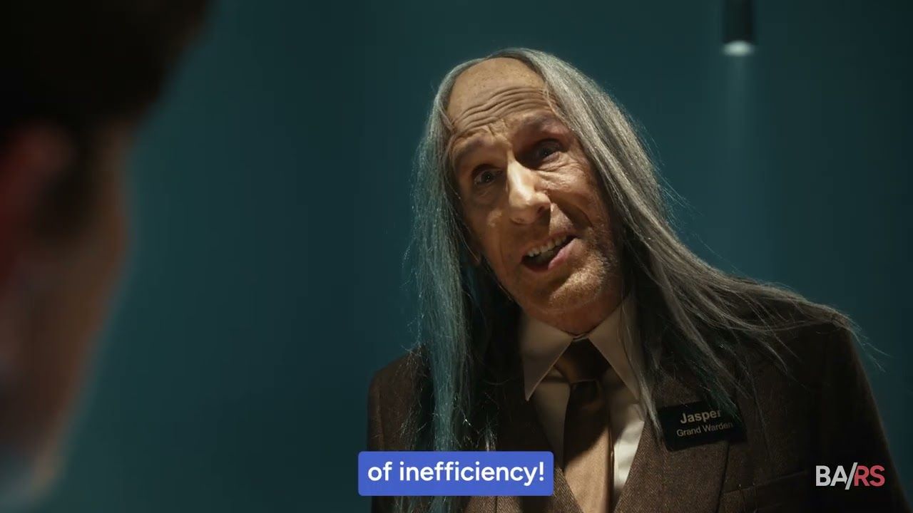Close-up of an older man with long gray hair in a suit and name badge, speaking against a teal background.