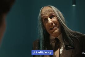 Close-up of an older man with long gray hair in a suit and name badge, speaking against a teal background.