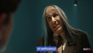 Close-up of an older man with long gray hair in a suit and name badge, speaking against a teal background.