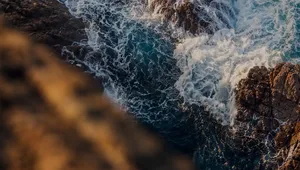 Waves crashing against rugged brown rocks with foamy white water in a vivid, turbulent sea.