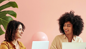 Two women smiling while working on laptops at a white desk, with a pink wall and green plant in the background.