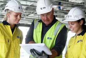 Three construction workers in safety helmets and yellow jackets review plans on a construction site.