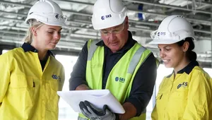Three construction workers in safety helmets and yellow jackets review plans on a construction site.