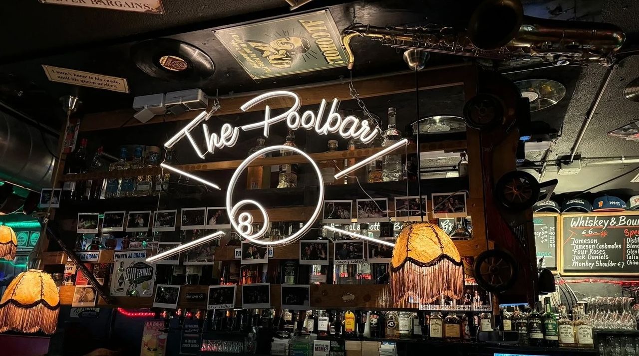Neon sign reading "The Poolbar" with hanging lamps, photos, and a saxophone in a cozy bar setting.