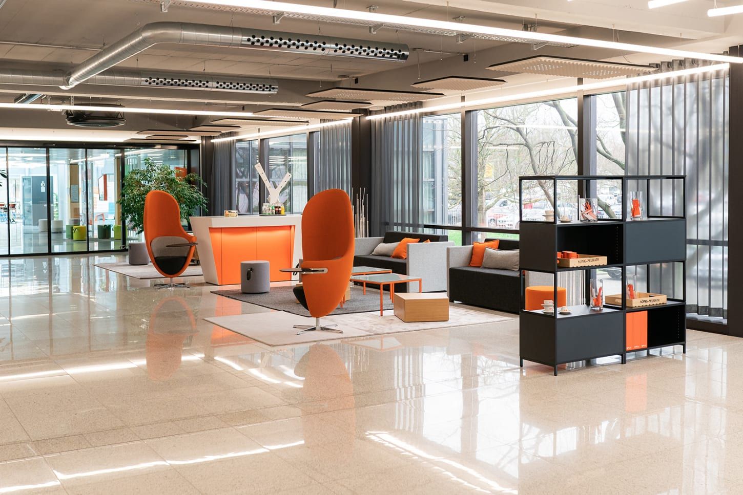 Modern office lobby with sleek furniture, orange accents, large windows, and a polished floor. Shelves with items are in the foreground.