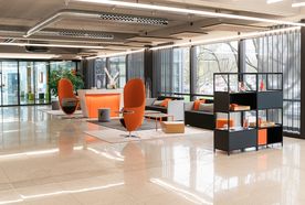 Modern office lobby with sleek furniture, orange accents, large windows, and a polished floor. Shelves with items are in the foreground.