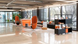 Modern office lobby with sleek furniture, orange accents, large windows, and a polished floor. Shelves with items are in the foreground.
