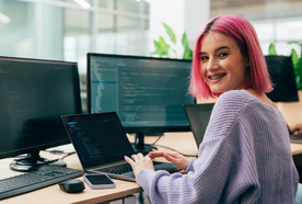 Person with pink hair smiling while working on a laptop in an office, surrounded by multiple computer screens displaying code.