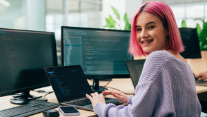 Person with pink hair smiling while working on a laptop in an office, surrounded by multiple computer screens displaying code.