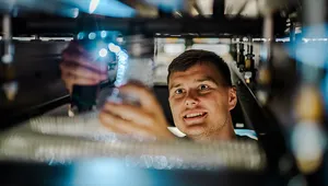 A technician smiles while inspecting industrial machinery from below, holding a small illuminated component amid cables, tubing, and metal equipment parts, with blue-toned lighting reflecting off surrounding surfaces.