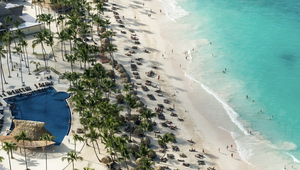 Aerial view of a tropical beach with turquoise water, palm trees, sun loungers, and a pool, with people enjoying the sun and sea.