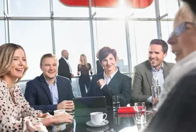 A group of people in business attire sitting around a table in a bright office, smiling and engaged in conversation.
