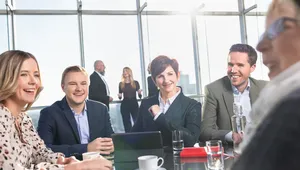 A group of people in business attire sitting around a table in a bright office, smiling and engaged in conversation.