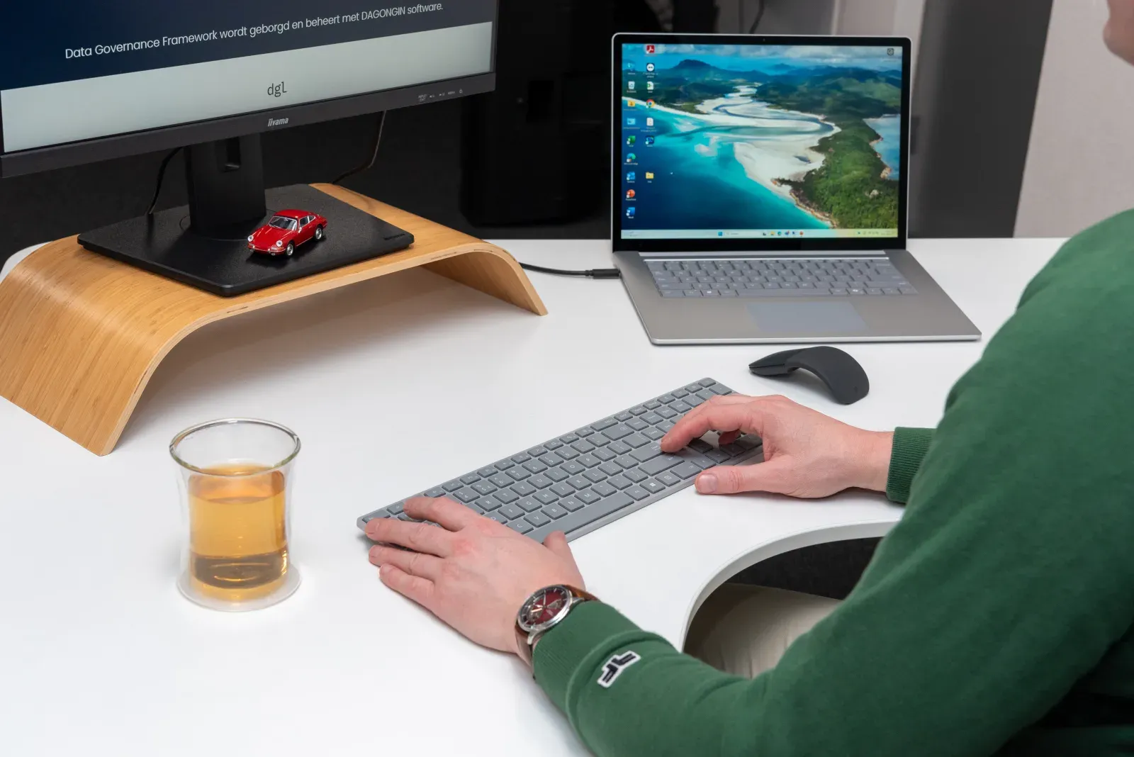 Person typing on a keyboard at a desk with a monitor and laptop displaying various screens. A glass of tea and small red car model sit nearby.