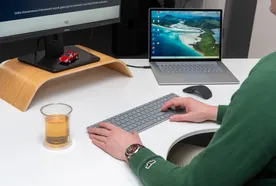 Person typing on a keyboard at a desk with a monitor and laptop displaying various screens. A glass of tea and small red car model sit nearby.