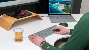 Person typing on a keyboard at a desk with a monitor and laptop displaying various screens. A glass of tea and small red car model sit nearby.