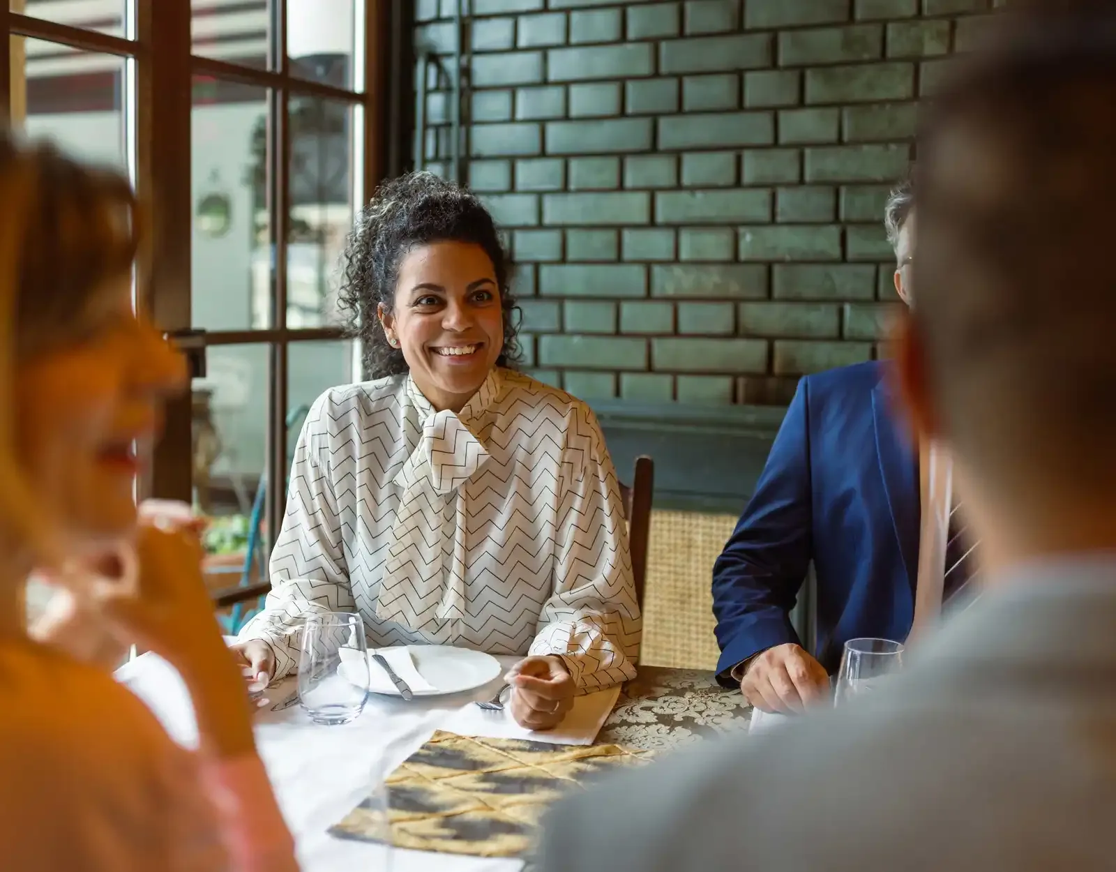 A woman in a patterned blouse sits at a table, smiling while dining with others in a warmly lit restaurant.