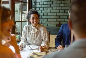 A woman in a patterned blouse sits at a table, smiling while dining with others in a warmly lit restaurant.