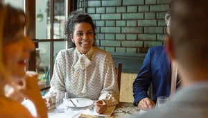 A woman in a patterned blouse sits at a table, smiling while dining with others in a warmly lit restaurant.