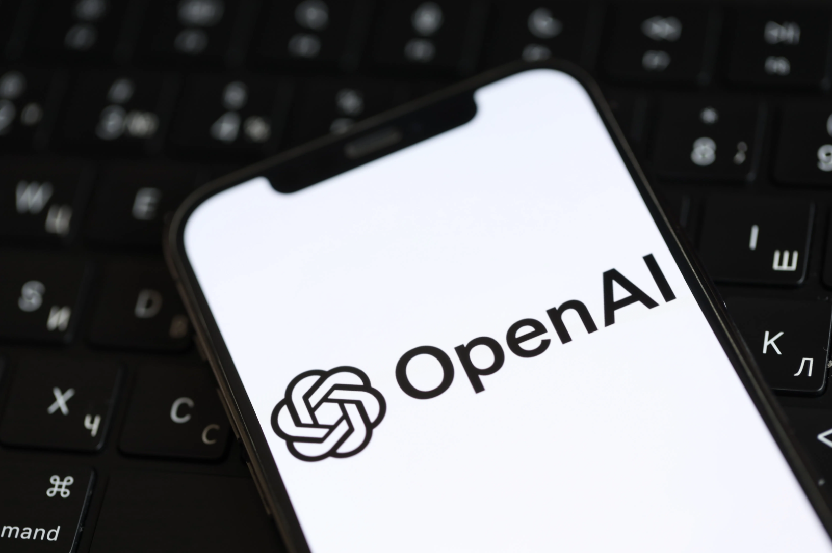 A smartphone displaying the OpenAI logo and text, resting on a keyboard with Cyrillic and Latin characters.