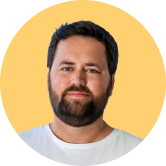 A bearded man with short dark hair is wearing a white shirt against a yellow circular background.