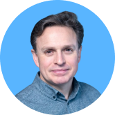 A man with short, dark hair wearing a blue button-up shirt poses against a solid blue background.