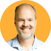 Headshot of Deane Barker. Smiling man with a beard and short hair, wearing a light blue shirt, against an orange background