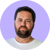 A bearded man with short hair and a white shirt is looking forward, set against a light purple background.