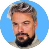 Man with silver hair and a beard, wearing a turtleneck sweater, against a blue background.