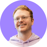 Smiling person with glasses and red hair, wearing a lavender hoodie, against a light purple circular background.