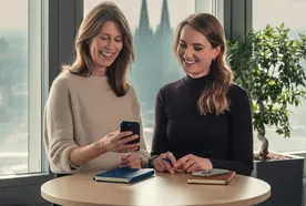Two women smiling and looking at a smartphone over a round table, with notebooks, in a room with large windows and a view of a cathedral.