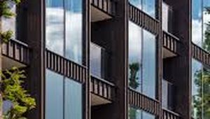 Modern apartment building with reflective glass windows and brown exterior, surrounded by green trees under a clear blue sky.