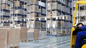 A worker in blue overalls and a yellow helmet operates a forklift in a large warehouse filled with stacked pallets and shelves.