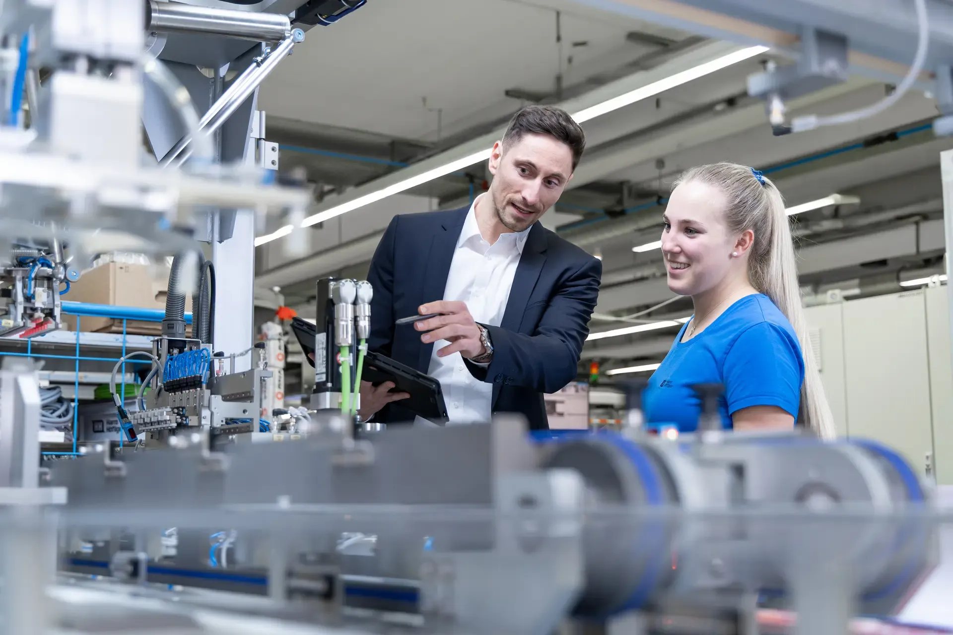 A man in a business suit and a young woman in a blue work shirt stand together in a modern industrial facility. The man holds a tablet and points toward a piece of machinery while explaining something, and the woman smiles as she listens. Various mechanical components and production equipment are visible in the foreground and background.