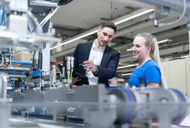 A man in a business suit and a young woman in a blue work shirt stand together in a modern industrial facility. The man holds a tablet and points toward a piece of machinery while explaining something, and the woman smiles as she listens. Various mechanical components and production equipment are visible in the foreground and background.