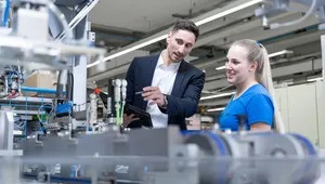 A man in a business suit and a young woman in a blue work shirt stand together in a modern industrial facility. The man holds a tablet and points toward a piece of machinery while explaining something, and the woman smiles as she listens. Various mechanical components and production equipment are visible in the foreground and background.
