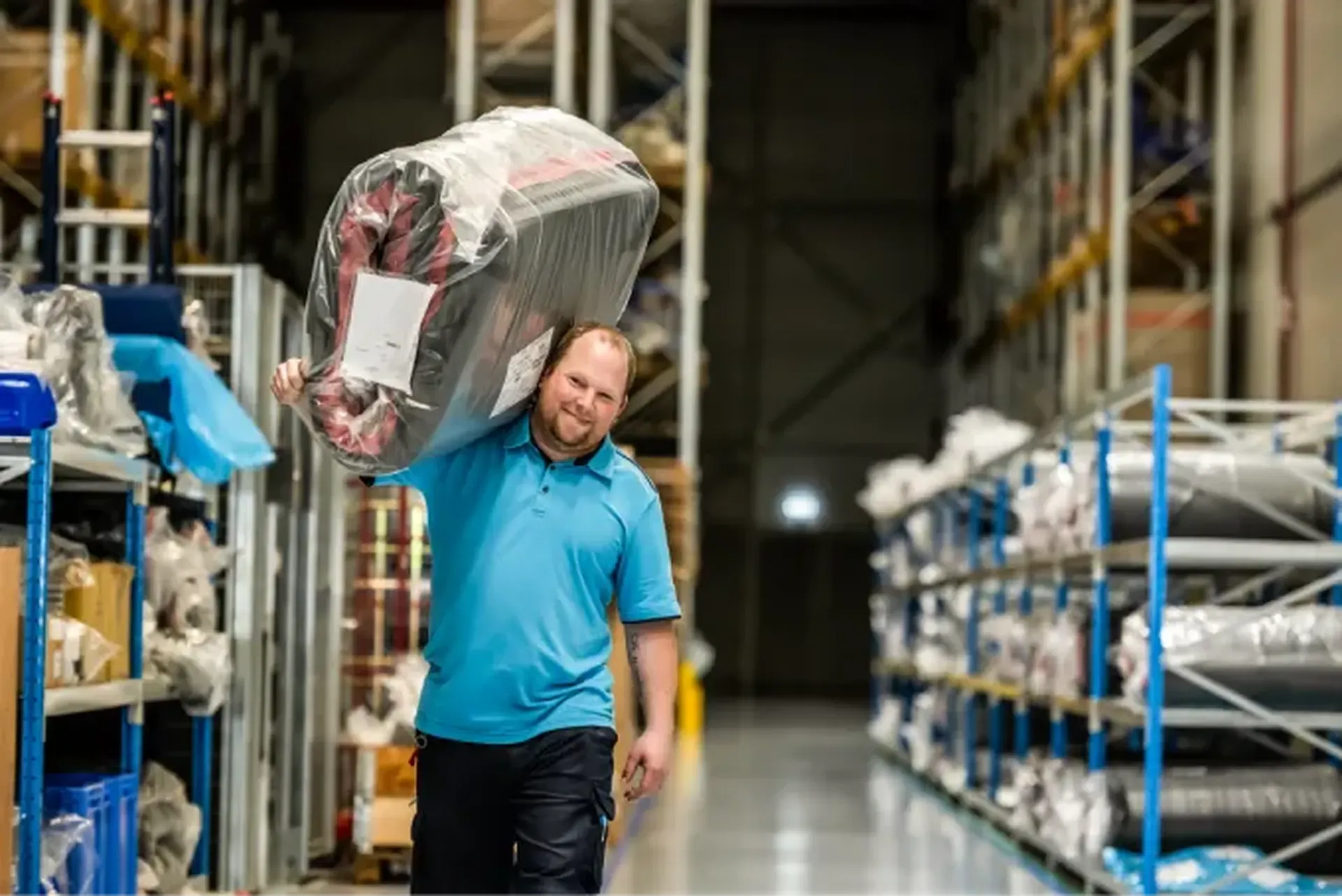 Man in a blue polo shirt carrying a large, wrapped package on his shoulder in a warehouse aisle with shelves and various items.